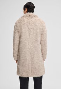 Persona con capelli scuri e corti che indossa un lungo cappotto di pelliccia beige chiaro visto da dietro su uno sfondo semplice.