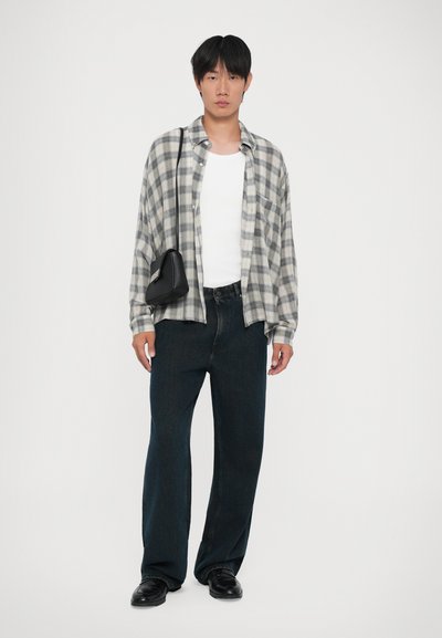 Άτομο που φοράει ένα γκρι καρό πουκάμισο πάνω από ένα λευκό κοντομάνικο μπλουζάκι, σκούρα φαρδιά τζιν, μαύρα παπούτσια και μια μαύρη τσάντα ώμου, στέκεται μπροστά σε λευκό φόντο.