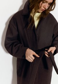 Mørkebrun uldfrakke med en stor krave, oversized pasform, knapper og et tøjret bælte; båret over en lysegul sweater.