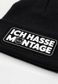 Black beanie hat with ribbed texture, featuring a rectangular white patch that reads "ICH HASSE MONTAGE" with a black cat graphic.