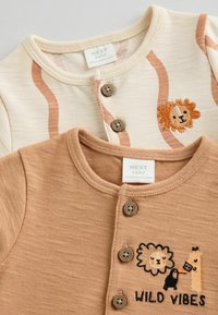 Två babytröjor, en krämfärgad med orange ränder och lejonbroderi, den andra beige med texten "WILD VIBES" och knappar. Mjuk tyg, rund hals.