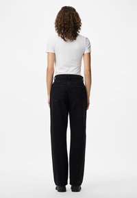 Person med krøllete hår iført en enkel hvit t-skjorte og høytlivs svarte jeans som står med ryggen mot på en ensfarget hvit bakgrunn.