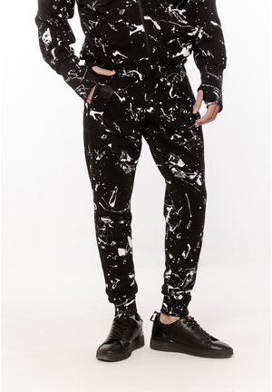 Osoba ubrana w czarne dresy i kurtkę z białym wzorem rozbryzganej farby, do tego czarne niskie trampki, stojąca z rękami w kieszeniach.