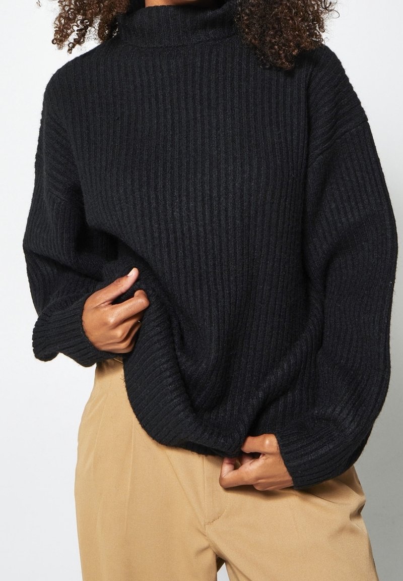 Svart ribbad polotröja med lös passform, utrustad med långa ärmar och ett texturerat stickat mönster. Tillsammans med beige byxor.