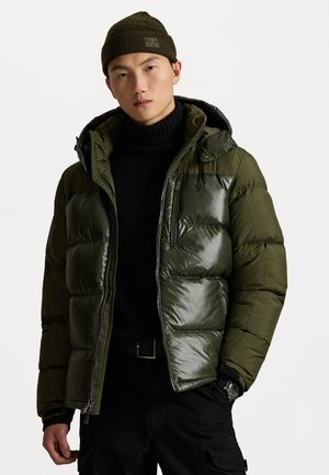 Dunkelgrüne Pufferjacke mit glänzenden und matten Abschnitten, Kapuze, Reißverschlusstaschen und genähten Details. Über einem schwarzen Rollkragenpullover getragen.