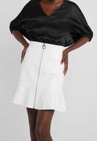 Personne à la peau foncée portant un chemisier en satin noir à manches bouffantes et une jupe blanche taille haute avec une fermeture éclair à l'avant et un ourlet évasé.