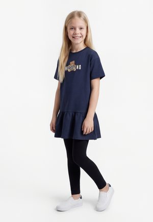 Smilende jente med langt blondt hår iført en marineblå kjole med teddybjørnlogo, svarte leggings og hvite slip-on sko, stående mot en hvit bakgrunn.