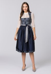 Navyblaues Dirndl mit besticktem Mieder, Satinsrock und grauem Band zum Binden in der Taille. Langärmliges Spitzenshirt sorgt für Textur und Kontrast.