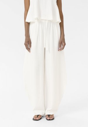 Personne portant un pantalon blanc ample texturé avec une taille à cordon et un haut sans manches ample assorti, des sandales noires à lanières visibles.