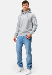Sudadera gris con capucha ajustable, puños y dobladillo acanalados. Combinada con unos vaqueros azul claro y zapatillas deportivas blancas, de pie frente a un fondo simple.