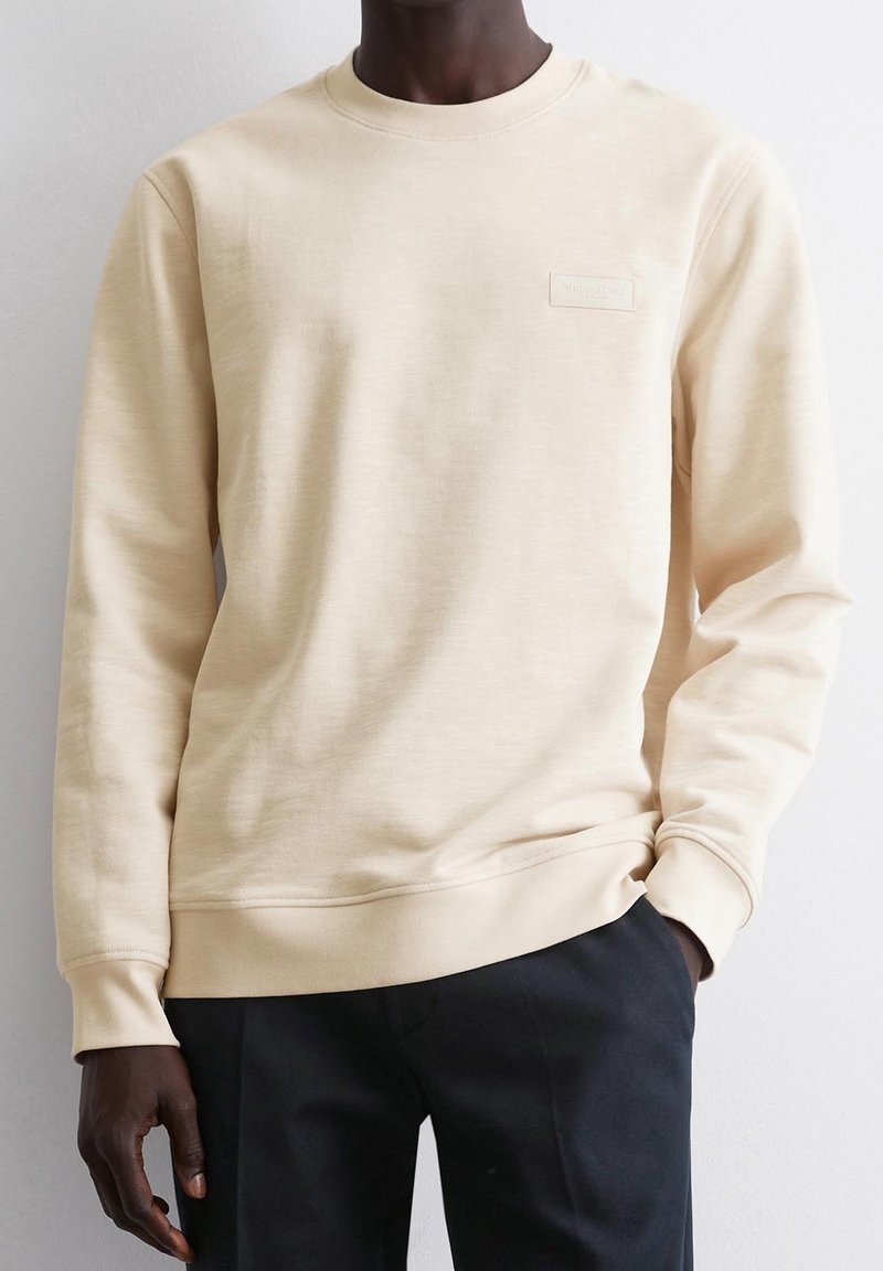 Homme portant un sweat-shirt beige clair à manches longues et un pantalon noir, une main dans la poche, debout contre un mur uni.
