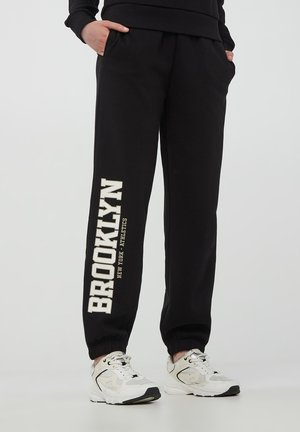 Pantaloni tuta neri in morbido tessuto con fascette elastiche; presentano un grande testo bianco "BROOKLYN" e una stampa più piccola sulla gamba sinistra.