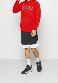 Sudadera roja con capucha con el texto "ANGELS", pantalones cortos deportivos negros con ribete blanco y zapatillas deportivas negras. El modelo se encuentra en un fondo neutro.