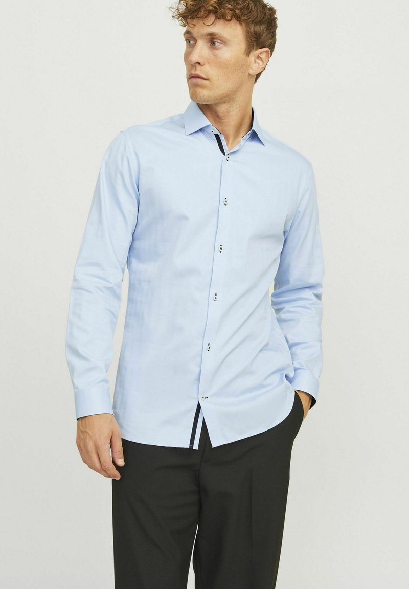 Chemise à manches longues bleu clair, design à boutons, texture subtile, accents noirs contrastants sur les boutons et l'ourlet, coupe ajustée, associée à un pantalon noir.