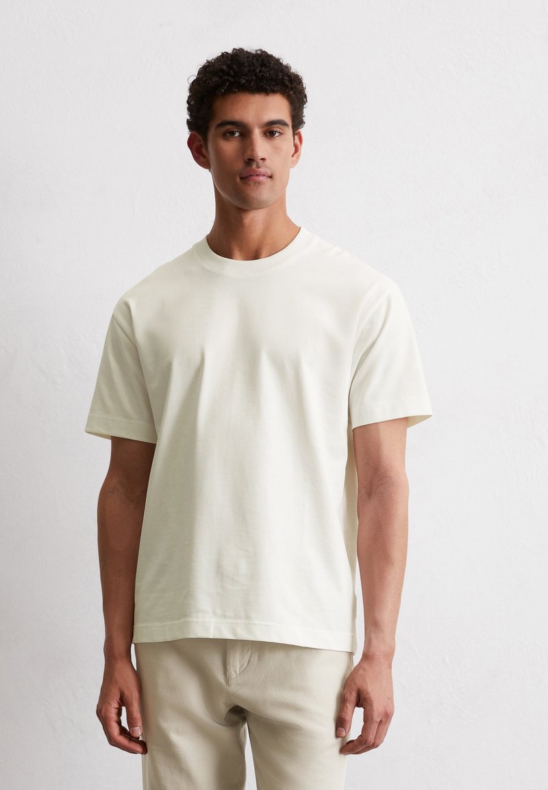 T-shirt beige clair à manches courtes et col rond en tissu coton lisse, avec une coupe décontractée et des coutures nettes. Porté avec un pantalon clair.