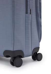 Modrá látková cestovní taška s texturovaným povrchem, sippovým uzávěrem, boční kapsou a černými kolečky. Design je obdélníkový s zaoblenými rohy.