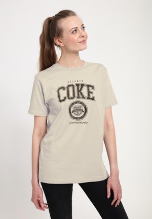 Světle béžové tričko s černou grafikou "ATLANTA COKE", obsahující kruhové logo Coca-Coly a slogan. Krátké rukávy, pohodlný střih.