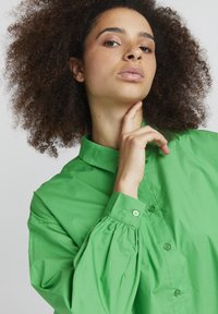 Green cotton shirt with a stand collar, button front, and puffed sleeves. Smooth texture with subtle sheen and matching buttons.