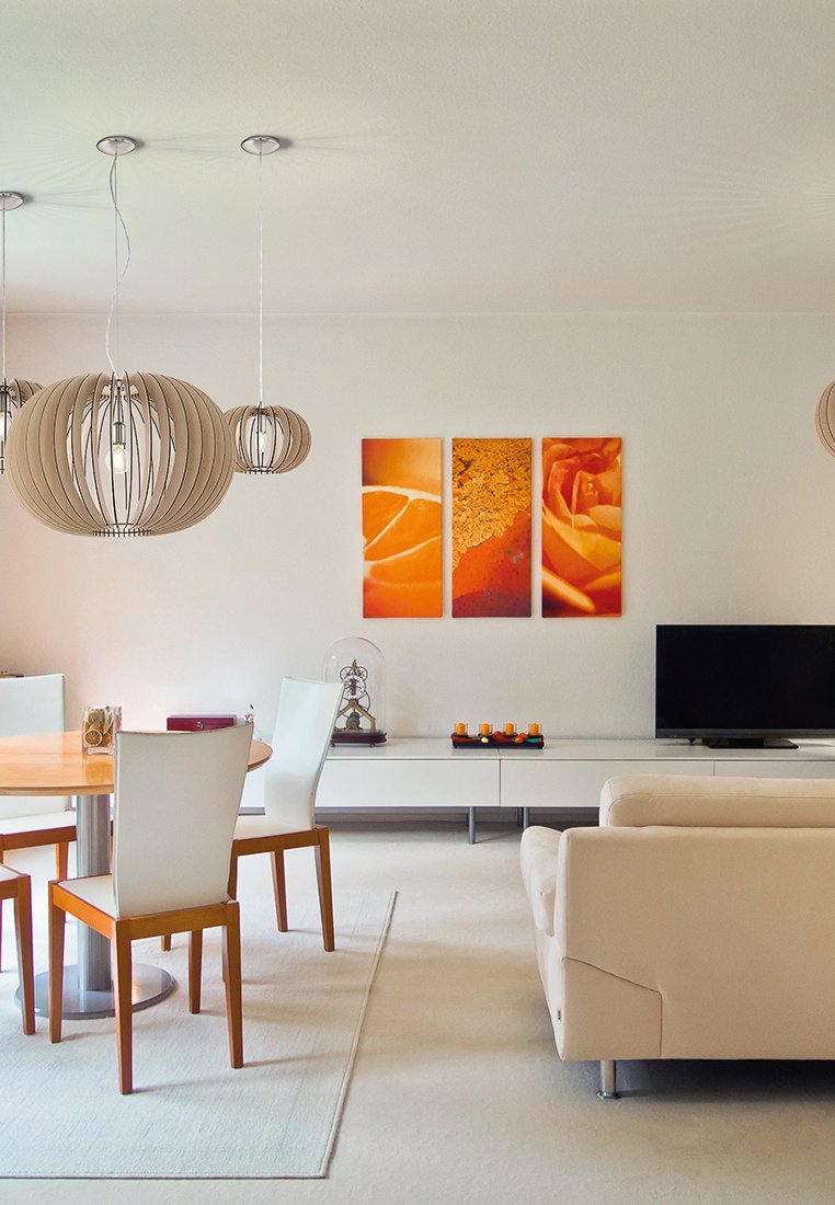 Offener Wohnbereich mit einem hellen Holztisch, weißen Stühlen, einem beigen Sofa und dekorativer orangefarbener Wandkunst. Pendelleuchten hängen darüber.
