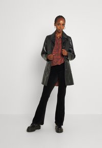 Glanzende donkergroene waterdichte trenchcoat met een kraag, knoopsluiting en zijzakken, gedragen over een rood geruite shirt en zwarte corduroy broek.