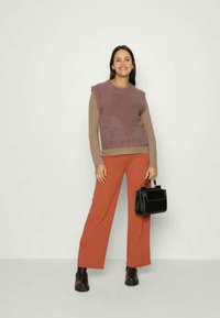 Top roxo de malha sem mangas sobre uma camisa bege de mangas longas, combinado com umas calças largas laranja e botas pretas até ao tornozelo, segurando uma pequena bolsa preta.
