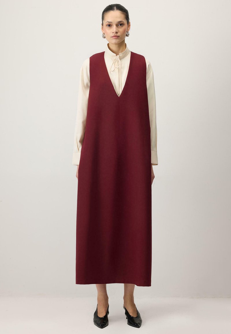 Vinrød ærmeløs kjole med dyb V-hals, lavet af glat stof, falder under knæene, kombineret med en cremefarvet bluse og sorte sko.