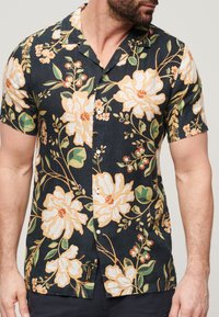 Camicia a maniche corte con sfondo nero e grandi motivi floreali bianchi e gialli, foglie verdi e chiusura con bottoni.