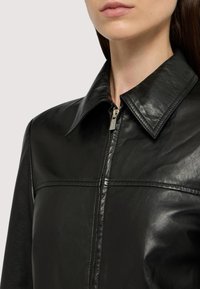 Černá kožená bunda se širokým límcem, zipovým zapínáním a viditelným švem. Povrch je hladký s mírným leskem.