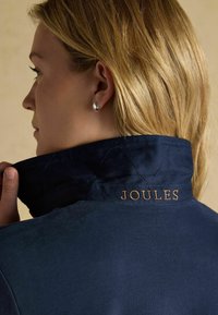 Marineblauer, gesteppten Kragen mit "JOULES" in Gold gestickt, Modell hält den Kragen mit glatter Textur. Silberohrringe sind im Ohr sichtbar.