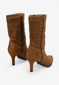Botines de ante marrón con detalles de tachuelas plateadas, cremallera lateral y un tacón alto y delgado. Superficie texturizada con un interior suave.