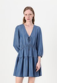 Denim kleita ar V-veida izgriezumu un šņuķīšu detaļu. Iespējota garas pūkainas piedurknes, savāktā josta un izplestais, slāņotais svārks gaismas zilā krāsā.