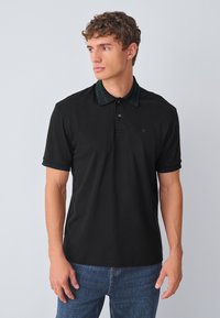 Juoda polo marškinėliai su žaliai juodu languotu apykakle, trumpomis rankovėmis ir subtiliu logotipu krūtinėje, dėvimi su mėlynomis džinsais.