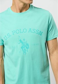 Tricou din bumbac turcoaz, cu guler rotund, având un imprimeu grafic cu jucători de polo și textul "U.S. POLO ASSN.", cu o textură netedă.