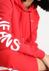 Felpa rossa con cappuccio e stampa bianca "JEANS" sulle maniche, dotata di cappuccio con coulisse e polsini a costine. Tessuto morbido in misto cotone.