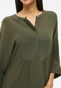 Blouse vert olive à coupe décontractée, avec patte de boutonnage et design plissé. Fabriquée en tissu texturé, elle dispose de manches trois-quarts.