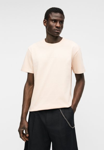 Άνδρας με σκούρο δέρμα που φοράει ένα ανοιχτό μπεζ κοντομάνικο μπλουζάκι και μαύρο παντελόνι με ασημένια αλυσίδα, στέκεται μπροστά σε ένα απλό φωτεινό φόντο.