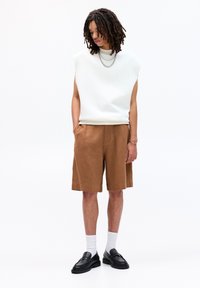 Hvid ærmeløs top med rund hals, parret med brune knæ-lange shorts og sorte loafers. Modellen bærer sorte og hvide sokker samt en metallisk kæde.