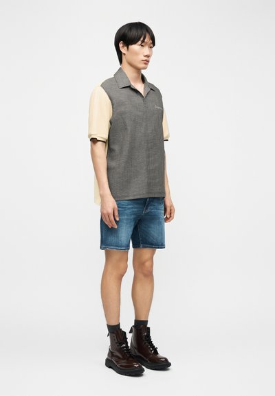 Άνδρας που φοράει διχρωμία κοντομάνικο πόλο πουκάμισο, τζιν σορτς, γκρι κάλτσες και σκούρα καφέ δερμάτινα μποτάκια με κορδόνια, στέκεται μπροστά σε απλό φόντο.