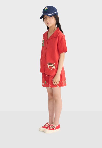 Ragazza con maglietta rossa a maniche corte e pantaloncini abbinati con stampe di animali da fattoria, cappellino da baseball blu scuro e scarpe rosse con fibbia, in piedi di profilo.
