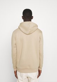 Muž otočený zády, oblečený v béžové mikině a off-white kalhotách na jednobarevném bílém pozadí.