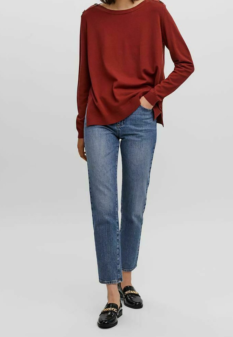 Maglietta rossa a maniche lunghe con scollo ampio, abbinata a jeans dritti blu chiaro, con vestibilità classica e mocassini neri con dettagli dorati.
