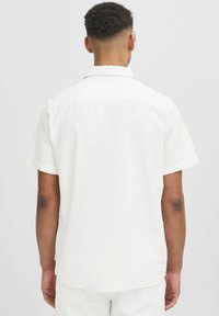 Camisa blanca de lino de manga corta con un corte relajado, yugo trasero y dobladillo curvado. Presenta costuras mínimas y sin hardware prominente.