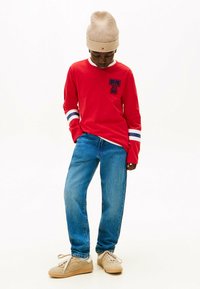 Camisa roja de mangas largas con rayas azules y blancas en las mangas, logo en el pecho; combinada con vaqueros de mezclilla azules y zapatillas deportivas beige. Gorro de punto beige.