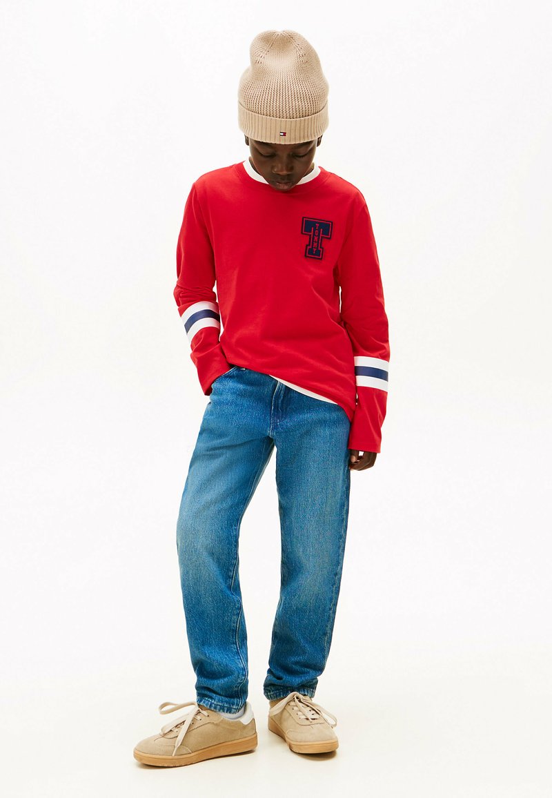 Camisa roja de mangas largas con rayas azules y blancas en las mangas, logo en el pecho; combinada con vaqueros de mezclilla azules y zapatillas deportivas beige. Gorro de punto beige.