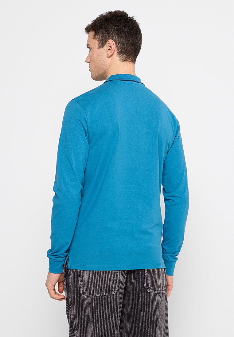 Polo a maniche lunghe in cotone blu brillante con colletto, caratterizzato da una texture morbida e un orlo dritto, abbinato a pantaloni a righe scure.