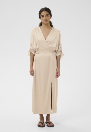 Kobieta ubrana w beżową jedwabną sukienkę kopertową z marszczonymi rękawami i przednim rozcięciem, stojąca boso na białym tle.