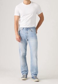 Homme portant un jean bleu clair, un t-shirt blanc à manches courtes et des baskets bleues, debout devant un fond uni.