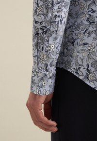 Lange mouwen shirt van lichtblauwe stof met een bloemenprint in donkerblauw. Knopsluiting bij de manchetten met witte knopen, die de verschillende patronen benadrukt.