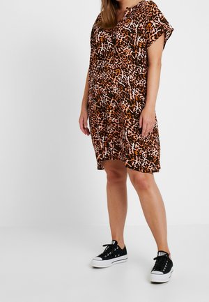 Femme portant une robe imprimée léopard longueur genoux et des baskets noires basses, debout devant un fond clair uni.