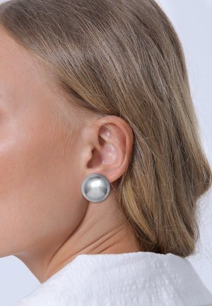Boucles d'oreilles circulaires en argent avec une surface lisse et réfléchissante, positionnées sur un fond clair, portées par une personne ayant des cheveux de longueur moyenne.